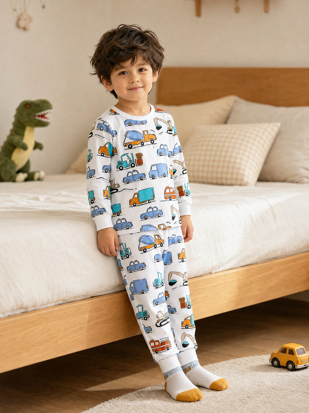 Cute boy wearing white city vehicle print long-sleeve cotton pajama set beside a wooden bed